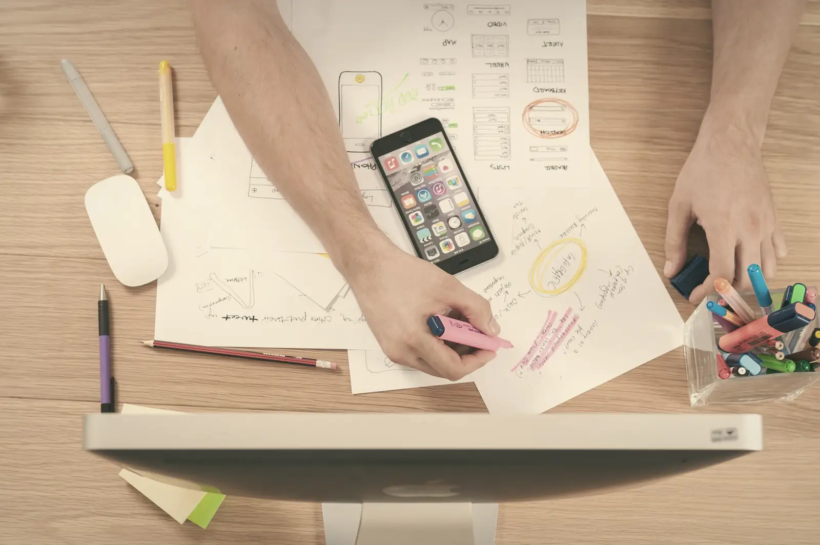 Hands marking up printed sketches and plans on a wooden desk.